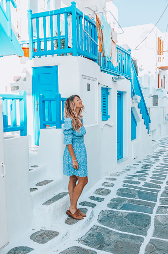 Portrait of a young beautiful happy traveler female lady in a blue dress enjoys the scenery while standing and looking up through the traditional white and blue small, whitewashed alleys of Mikonos town, Cyclades islands of Greece during sunny summer morning.