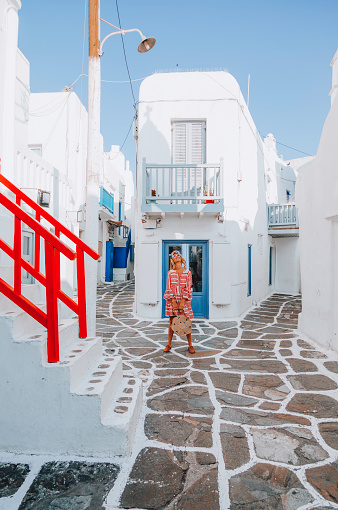 Portrait of a young beautiful happy traveler female lady in a red and white color dress and plastic frame sunglasses holding a mesh shopping bag posing and enjoying the scenery while standing and looking up through the traditional white and blue small, whitewashed alleys of Mikonos town, Cyclades islands of Greece during a sunny summer morning.