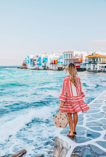 Back view of a young beautiful happy traveller female lady with a red dress and bag enjoys the scenery of city view landscape of The Little Venice district with old colourful houses by the sea coastline in Mykonos Town in Greek Islands at sunny day in Cyclades, Greece during summertime.