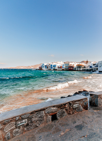 City view landscape of The Little Venice district from a beach with old colorful houses by the sea on sunny and windy summer day against blue sky in Mykonos Town in Greek Islands at sunset, Cyclades, Greece.
