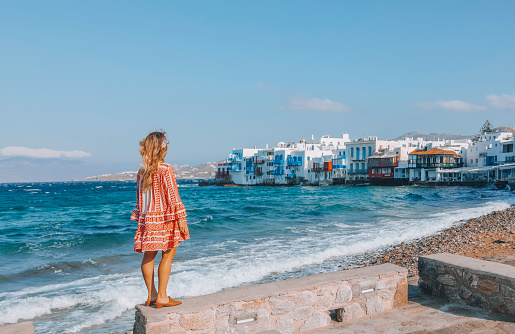 Rear view of a young beautiful happy traveler female lady with a red and white color dress and mesh shopping bag standing on a kerbside stone and enjoys looking the scenery of city view landscape of The Little Venice district with old colourful houses by the sea coastline in Mykonos Town in Greek Islands at sunny day in Cyclades, Greece during summertime.