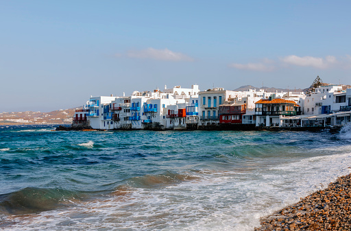 City view landscape of The Little Venice district from a beach with old colorful houses by the sea on sunny and windy summer day against blue sky in Mykonos Town in Greek Islands at sunset, Cyclades, Greece.