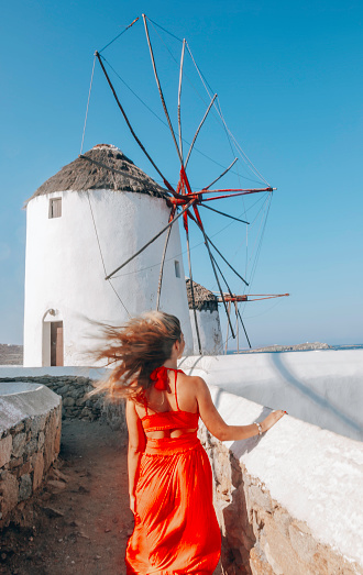 Young beautiful stylish happy female model tourist lady walking and enjoying the summer day view in a red color long dress looking at the view of famous traditional old white windmills in Mikonos town (Chora), Mykonos island, Cyclades, Greece on a sunny and windy summer day against blue clear sky. Travel Greece vacation luxury Europe cruise destination concept