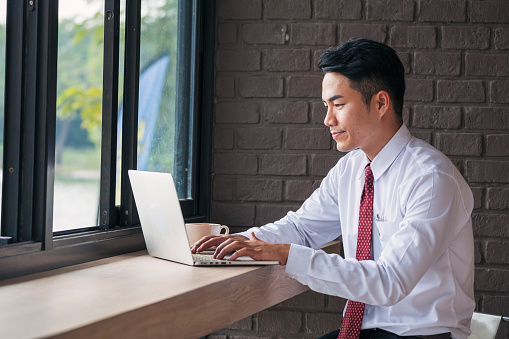 Portrait Asian Businessman typing laptop computer drinking coffee cup at office desk via wireless internet technology working online. Asian handsome man Freelance business person confident manager