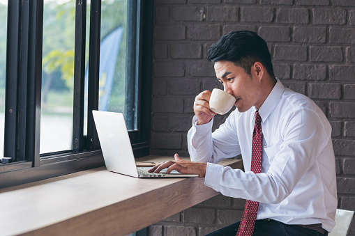 Portrait Asian Businessman typing laptop computer drinking coffee cup at office desk via wireless internet technology working online. Asian handsome man Freelance business person confident manager
