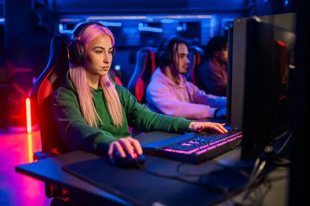Young woman with pink hair gamer playing PC video game and having fun with her friends. Female video game player participating on a tournament in a Esports arena or on a Gaming convention.