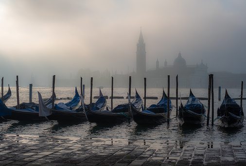 Gondolas and wooden posts in Grand Canal by pier with Church of San Giorgio Maggiore in the background against cloudy sky at Venice,Italy