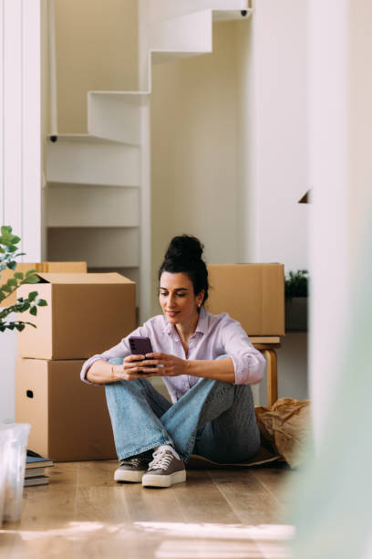 istock Woman Relaxing and Using Smartphone Amid Moving Boxes at Home 2115959766
