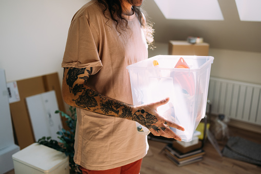 Adult with tattoos holds a transparent storage box in a sunlit room, suggesting organization and decluttering.