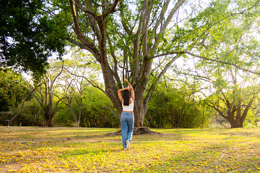rear or back view of pretty latin hispanic woman dressed casual in white and jeans stretching standing in front of tree in a park during golden hour