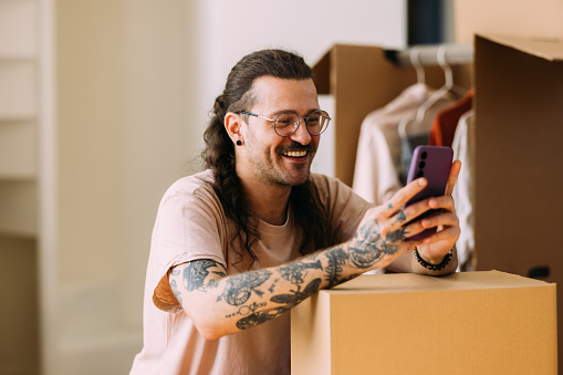 Joyful tattooed adult male with glasses using a phone beside moving boxes in a bright room.