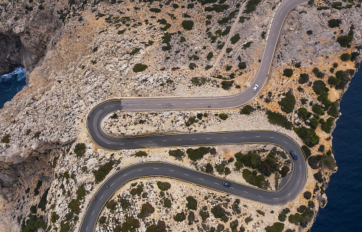 Lonely small white car driving by serpentine curved asphalt mountain road near Lighthouse of Cap de Formentor with seascape with rocky coast. Mallorca Island, Balearic Islands, Spain. Top aerial view.