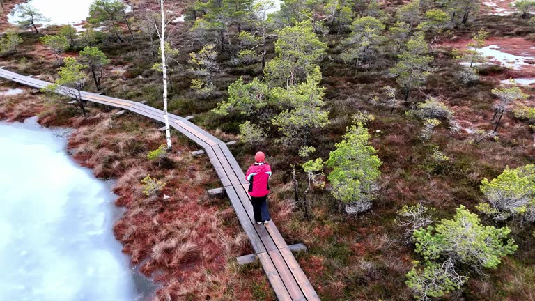 A woman is walking on a wooden bridge over a lake. The lake is frozen and surrounded by trees. The man is wearing a red jacket