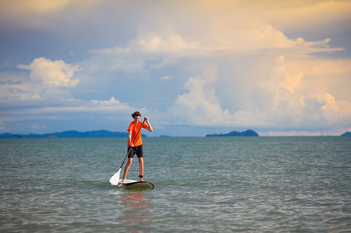 Young man on stand up paddle board. Water fun and beach sport. Healthy outdoor sports for summer vacation on tropical island. Holiday activity. Fit teenager boy training. Surfer exercising.