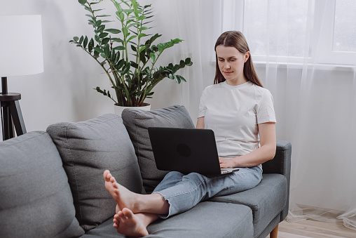 Pensive freelance young woman work on laptop settled down on couch in living room, looks thoughtful search ideas, correspond to client, solve business using wireless tech and internet connection
