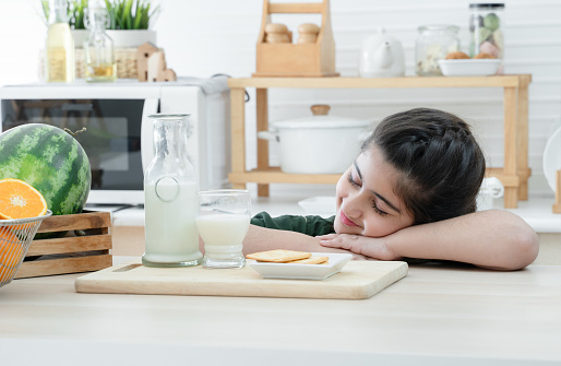 Cute Pakistani little Muslim child girl in traditional clothes sleeping at home kitchen. Fresh milk, Asian fruits in basket and crackers on table. Healthy breakfast food
