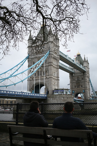 The two men seated on a park bench by the water, overlooking Tower Bridge.