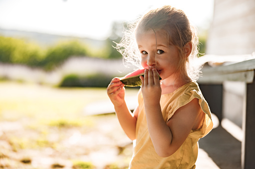 Cute little girl eating fresh watermelon in the backyard and looking at camera. Copy space.