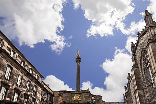 The historic Mercat Cross with its gleaming golden unicorn stands proudly in Parliament Square, Edinburgh, against a backdrop of ornate stone buildings and a picturesque blue sky with fluffy clouds