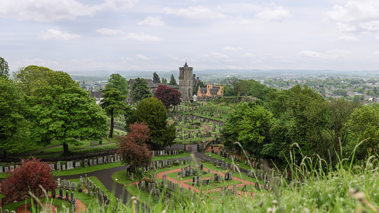 A sweeping panorama captures the serene Church of the Holy Rude Cemetery in Stirling, Scotland, nestled among lush foliage with the distant city beneath a soft sky