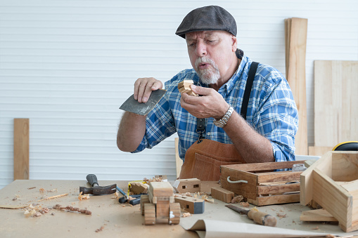 Caucasian senior old white bearded man carpenter in apron and hat working in workshop, use sandpaper polishing and blowing dust away from handmade wood toy, tools and handcrafts is on table