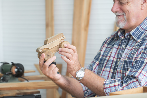 Selective focus on hands of Caucasian senior old white bearded man carpenter holding handmade wood car toy, smiling and looking at wooden toy after polished it. Carpentry working woodwork in workshop