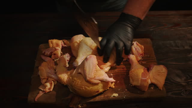 Man chopping chicken carcass with kitchen hatchet on cutting board. Male hands of cook cut raw chicken into pieces for cooking