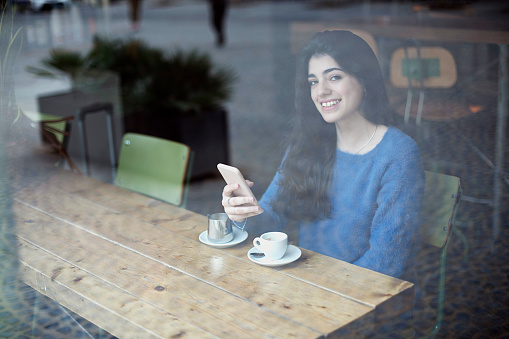 A young woman in a blue sweater focuses on her phone at a cafÃ©, with a coffee cup and saucer in front of her, viewed through a window