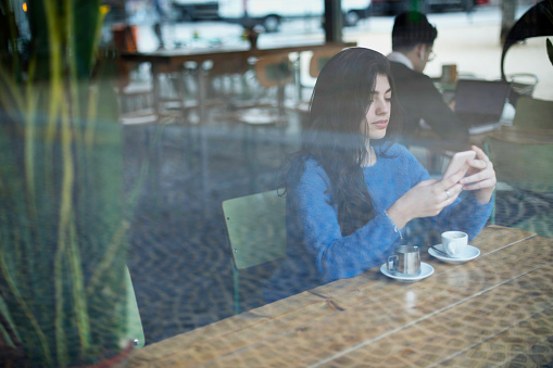A young woman in a blue sweater focuses on her phone at a cafÃ©, with a coffee cup and saucer in front of her, viewed through a window
