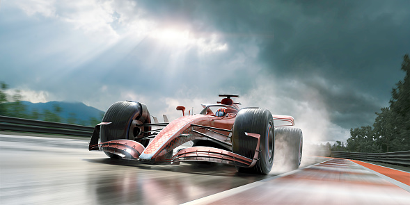 Low angle view of a generic red racing car with details driving at speed along a racetrack under wet conditions with water spray coming from wheels, under a stormy sky with bright sun peeking through clouds. With motion blur.