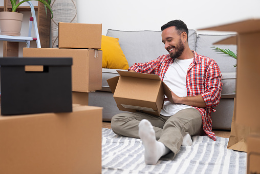 Brunette man sits near settee among boxes smilling in flat