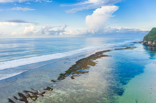 A coral reef stretches along the shoreline of a tropical beach, with clear blue waters on one side and lush greenery on the other. Waves gently crash against the reef, and in the distance, a rocky hill covered in green vegetation stands against a cloudy sky. Melasti beach, Bali.