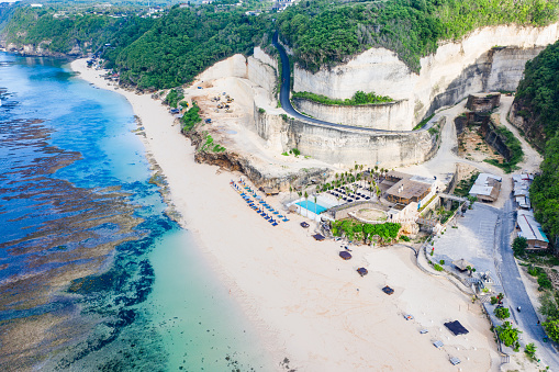 Aerial view of a tropical Melasti beach resort in Bali nestled against a lush green cliff. The crystal-clear turquoise waters reveal vibrant coral reefs. A winding road carved into the cliffside leads to the resort area, where blue umbrellas and lounging chairs await sunseekers.