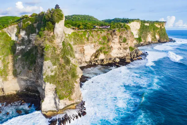 Aerial view reveals Uluwatu Temple, perched dramatically on the edge of a rocky cliff in Bali, Indonesia. The temple, surrounded by lush greenery, stands resilient against the elements. Below, turquoise sea waves relentlessly crash against the rugged rocks.
