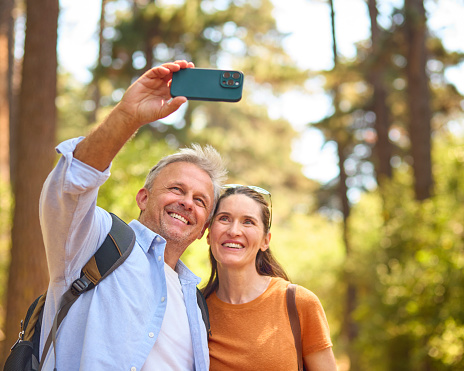 Retired Senior Couple Wearing Backpacks Hiking Through Countryside Posing For Selfie On Mobile Phone