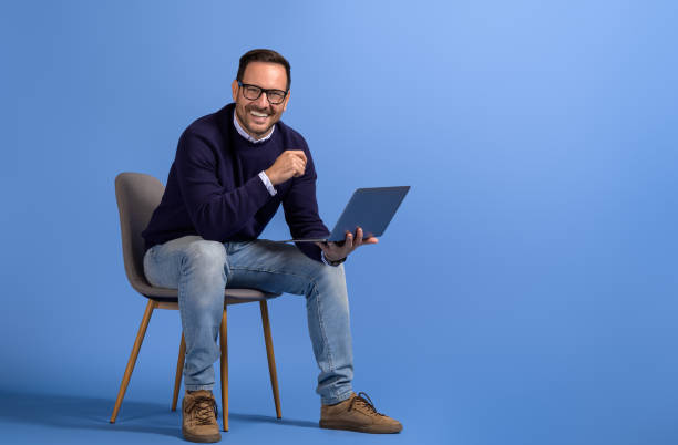 istock Successful male manager with laptop smiling at camera while sitting on chair over blue background 2116731282
