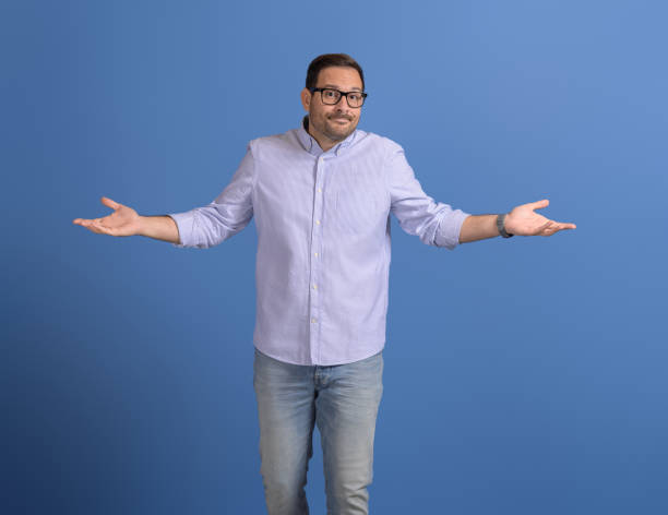 istock Confused businessman spreading hands and shrugging shoulders while standing against blue background 2116731297