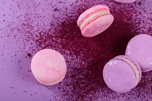 Pastel colored sweet french macaroons and splash of dry blueberry powder on purple background. Beautiful composition for bakery and pastry shop, top view