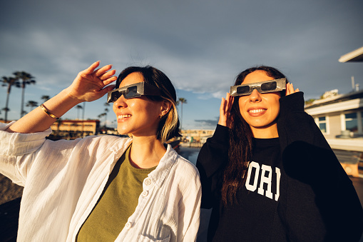 Two friends have fun together during a solar eclipse event. Their looking and pointing to the sun wearing the typical glasses normally used to watch a solar eclipse.