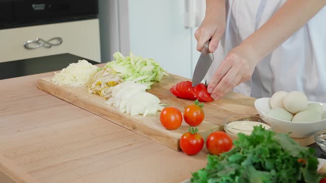The chef is preparing a frittata. Female hands with a sharp knife cut a tomato into slices, a wooden board, on a background of vegetables, diet food, healthy eating concept. Close-up, front view