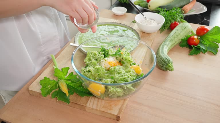 The chef is preparing zucchini. Beat eggs into a glass bowl with grated zucchini and season with salt, wooden table, vegetables in the background. Vegetarian food, zucchini pancakes, close-up, side view