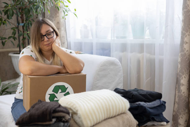 istock A woman is thoughtful with a box with a recycling sign in her hands while sorting out clothes. The woman was thinking about recycling her clothes or donating them to a charity. 2116801127