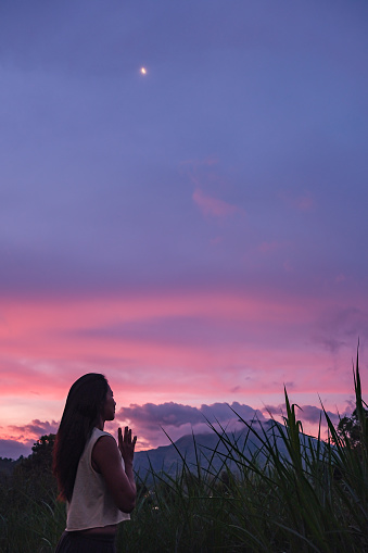 Vertical side shot of Asian Woman Doing Breathing Exercise in the Nature. She's standing alone with eyes closed and hands in prayer position. Her background is a mountain with beautiful sunset sky.