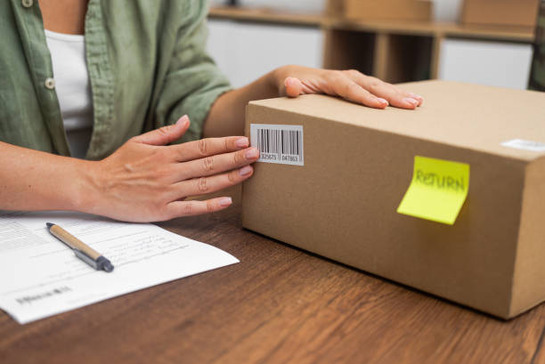 istock woman affixes a Return sticker, with a barcode, to cardboard box, online shopping refund process. 2116816214