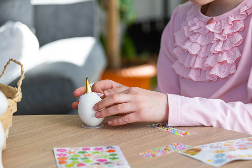 A cute girl with pink bunny ears makes an Easter craft - decorates an egg in the form of a unicorn with rhinestones, horn, flowers in the interior of a house with plants.