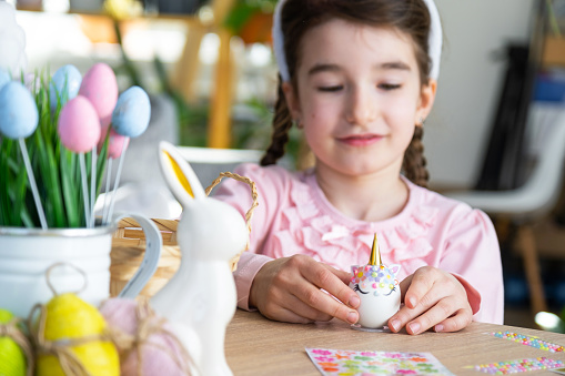 A cute girl with pink bunny ears makes an Easter craft - decorates an egg in the form of a unicorn with rhinestones, horn, flowers in the interior of a house with plants.