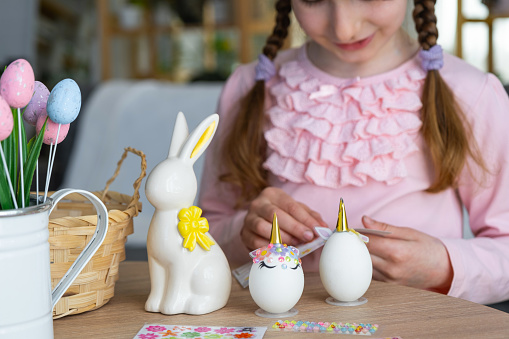 A cute girl with pink bunny ears makes an Easter craft - decorates an egg in the form of a unicorn with rhinestones, horn, flowers in the interior of a house with plants.