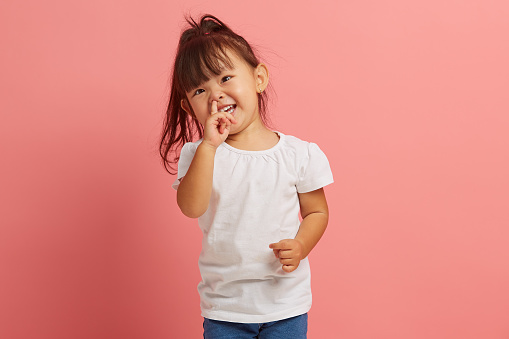 Studio portrait of little joyful Asian girl picking her nose on a pink isolated background with a free copy space. Capricious and stubborn child does not obey his parents. Concept of child crisis of three years old.