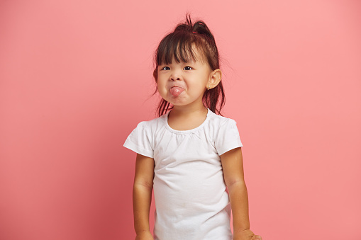 Studio portrait of little Asian baby girl shows tongue wears t shirt isolated over pink background. Three years old offended and capricious child.