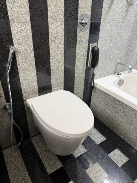 Stock photo showing a hotel bathroom featuring a white toilet with modern fittings include bathroom hygiene in the form of a hand shower bidet head attachment, which has been fixed to the wall next to the WC, complete with wall bracket and chrome hose connection. imagen de primer plano del baño del hotel de lujo, baldosas de pared y piso estampadas a rayas blancas y negras, inodoro de cerámica con botón pulsador colgado junto a la bañera, accesorio cromado para el cabezal del bidé de la ducha de mano, receptor